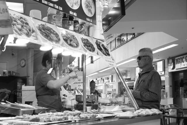 A black and white photo of a food court with a customer ordering from a vendor The vendor is behind the counter, and various food items are displayed on a menu above The customer is wearing sunglasses and a hat