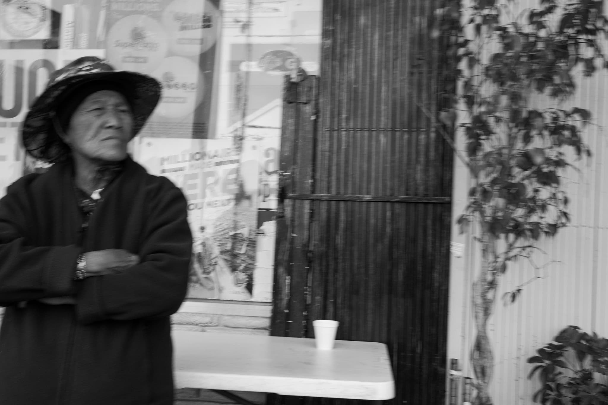 An elderly person wearing a hat and coat stands with arms crossed in front of a storefront, next to a table with a cup on it The background includes a wall with some climbing plants The image is in black and white