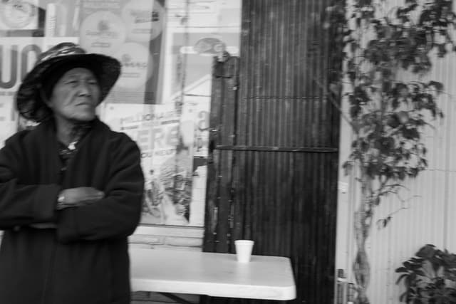 An elderly person wearing a hat and coat stands with arms crossed in front of a storefront, next to a table with a cup on it The background includes a wall with some climbing plants The image is in black and white