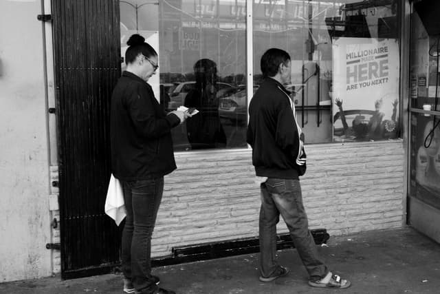 Two people standing outside a building, one looking at a phone and the other facing away, with a window and a sign in the background The scene is in black and white